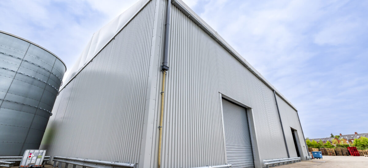 Large semi-permanent building with grey metal walls, grey doors, and a roller shutter door stands under a partly cloudy sky.