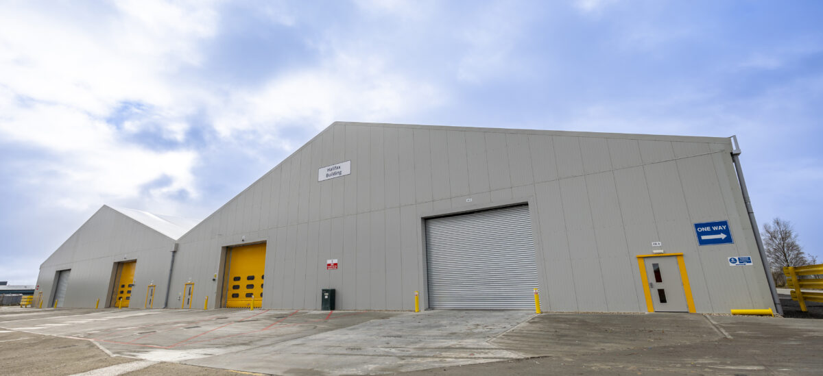 Large semi-permanent building with grey metal walls, yellow doors, and a roller shutter door stands under a partly cloudy sky.